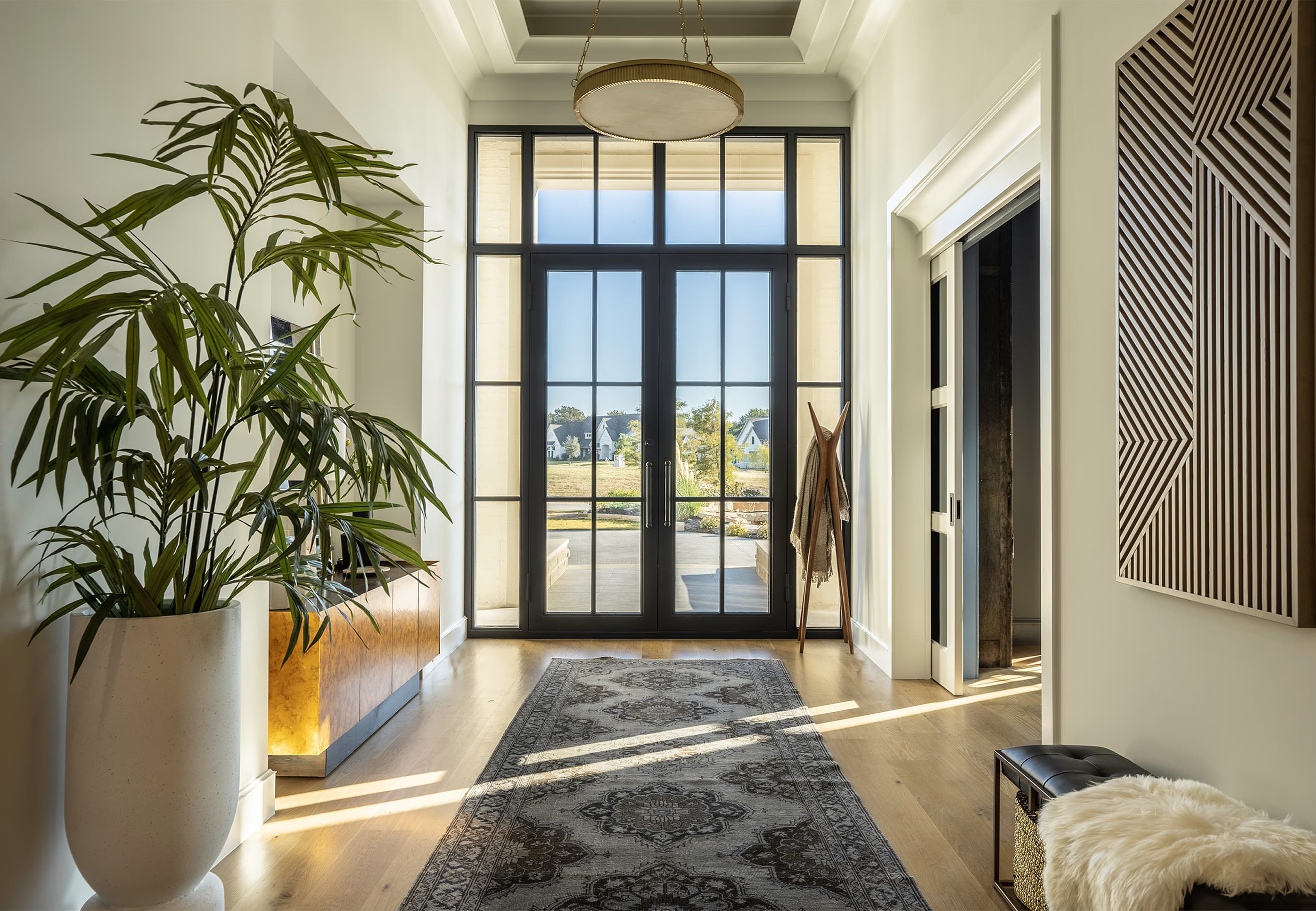 Wide pulled-back view of the entry: brass and alabaster chandelier overhead, vintage rug on white oak, French doors at center, geometric wood wall art at right