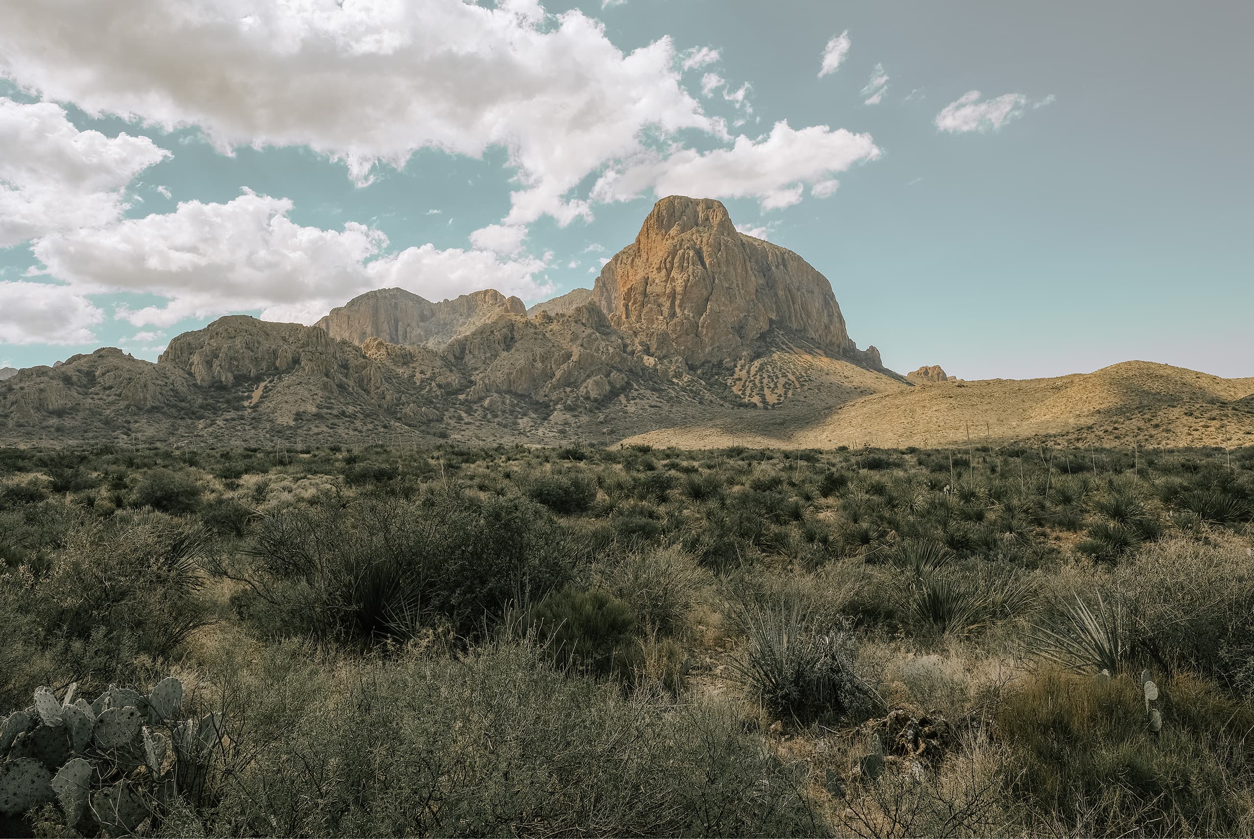 A lone peak in the Chisos Mountains rising over desert scrub and cactus, clouds stacked behind it