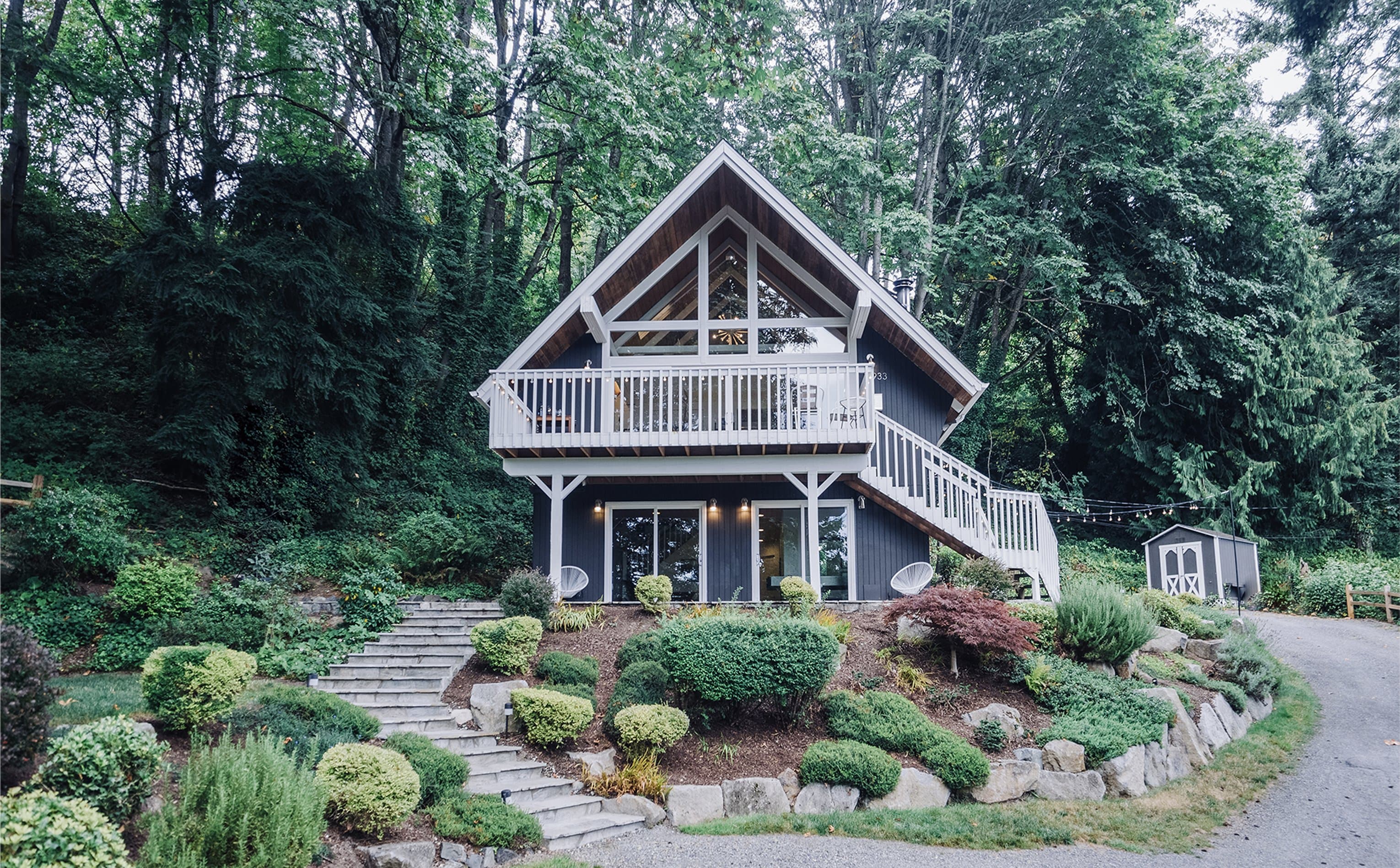 Front of the chalet repainted in warm gray with white railings, surrounded by Pacific Northwest evergreens and rocky landscape