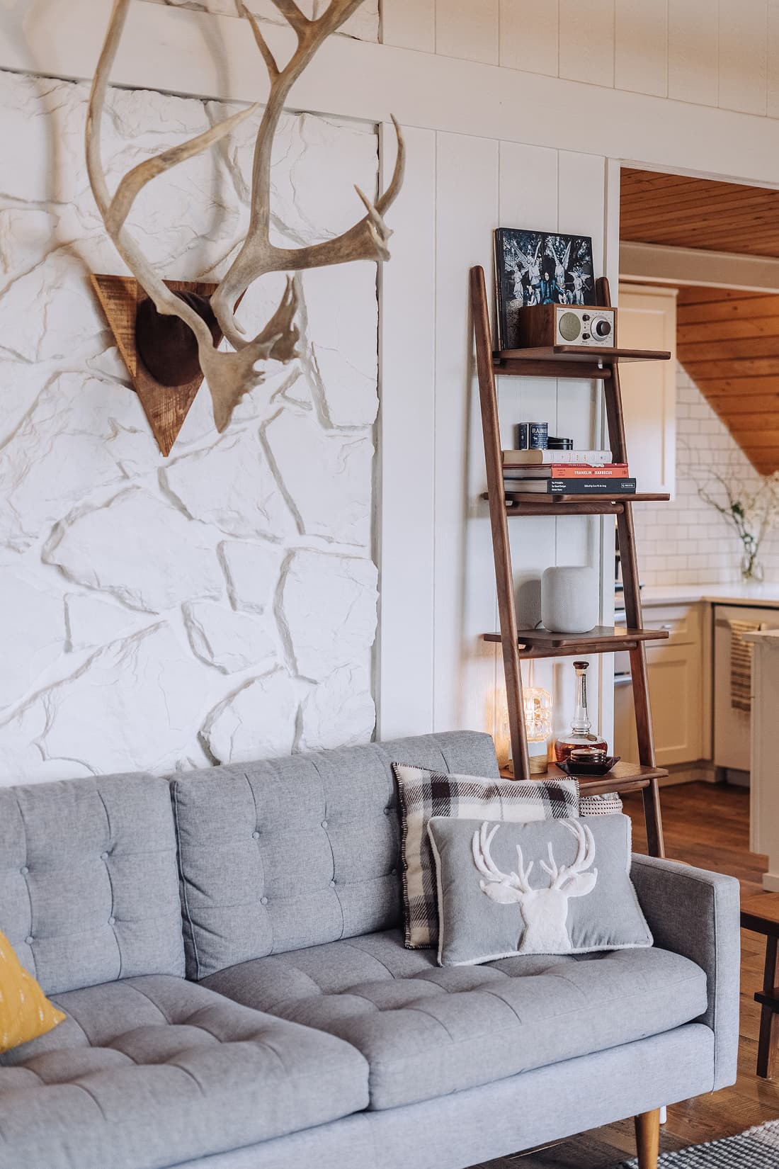 Closer detail of the tufted gray sofa with a stag-print pillow, walnut leaning ladder shelf with books and a speaker, against the painted stone wall