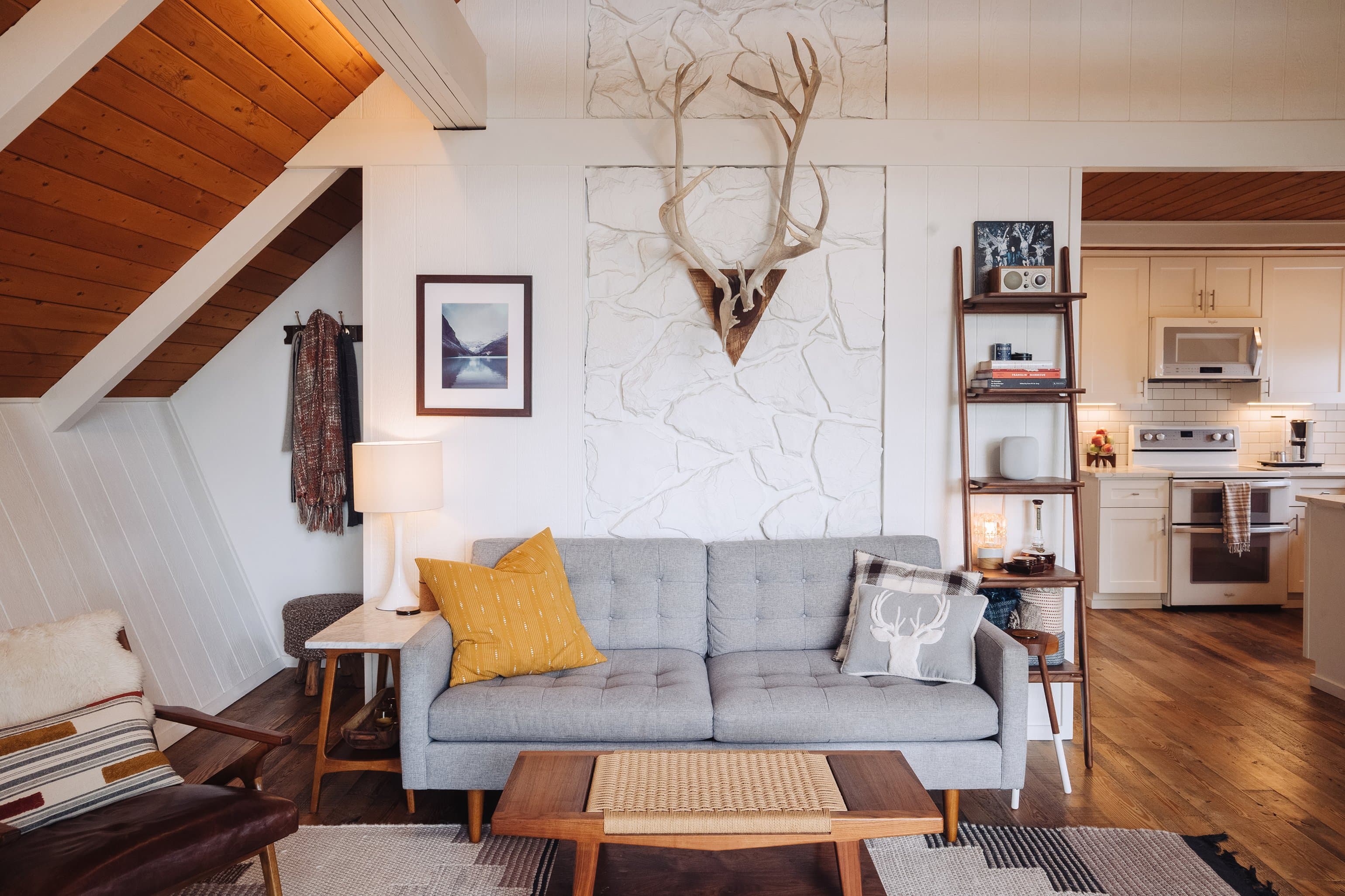 Chalet living area with tufted gray sofa, painted stone wall, antlers, walnut leaning ladder shelf, and the kitchen visible to the right