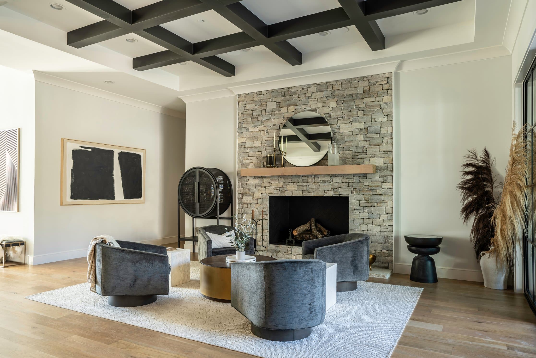 Pulled-back architectural view of the sitting room with black box beam coffered ceiling, stone fireplace, four swivel chairs around the brass coffee table, and pampas grass anchoring the corner