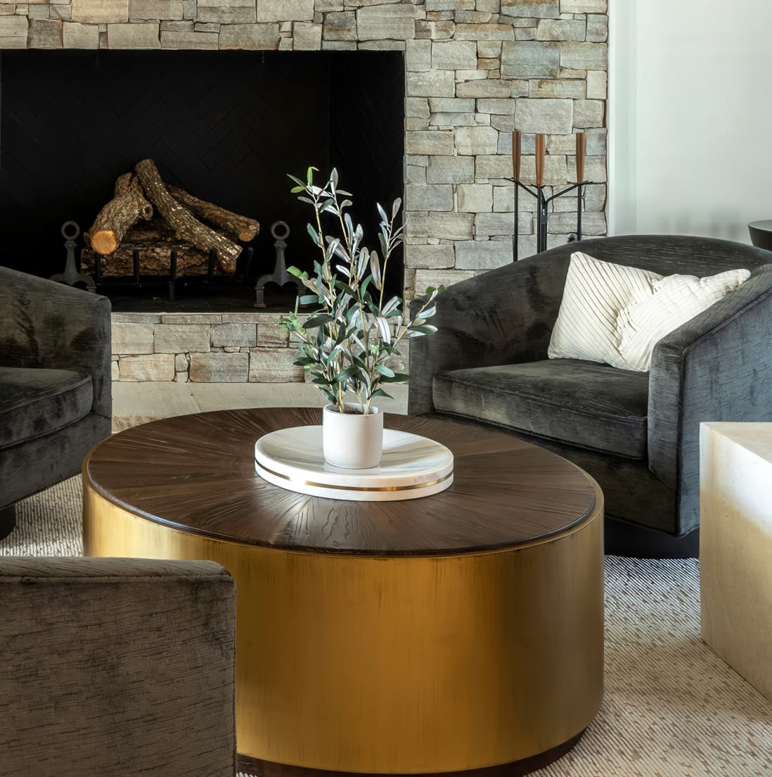 Tight detail of the round brass coffee table with wood top, olive plant in marble bowl, and the corner of a charcoal velvet swivel against the stone fireplace