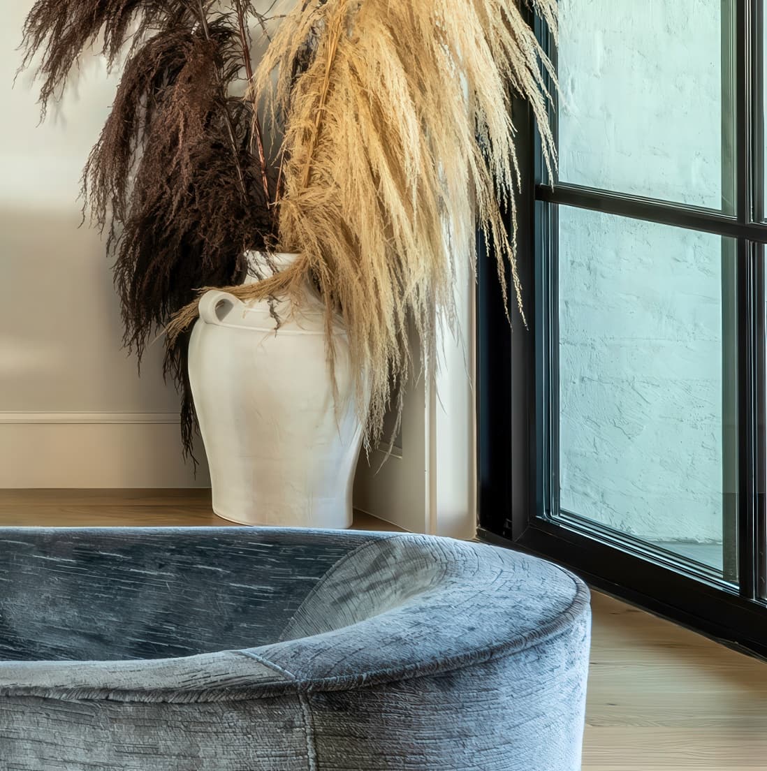 Pampas grass in a stone vase by a black-framed window with painted blue brick wall behind, edge of charcoal velvet swivel in foreground
