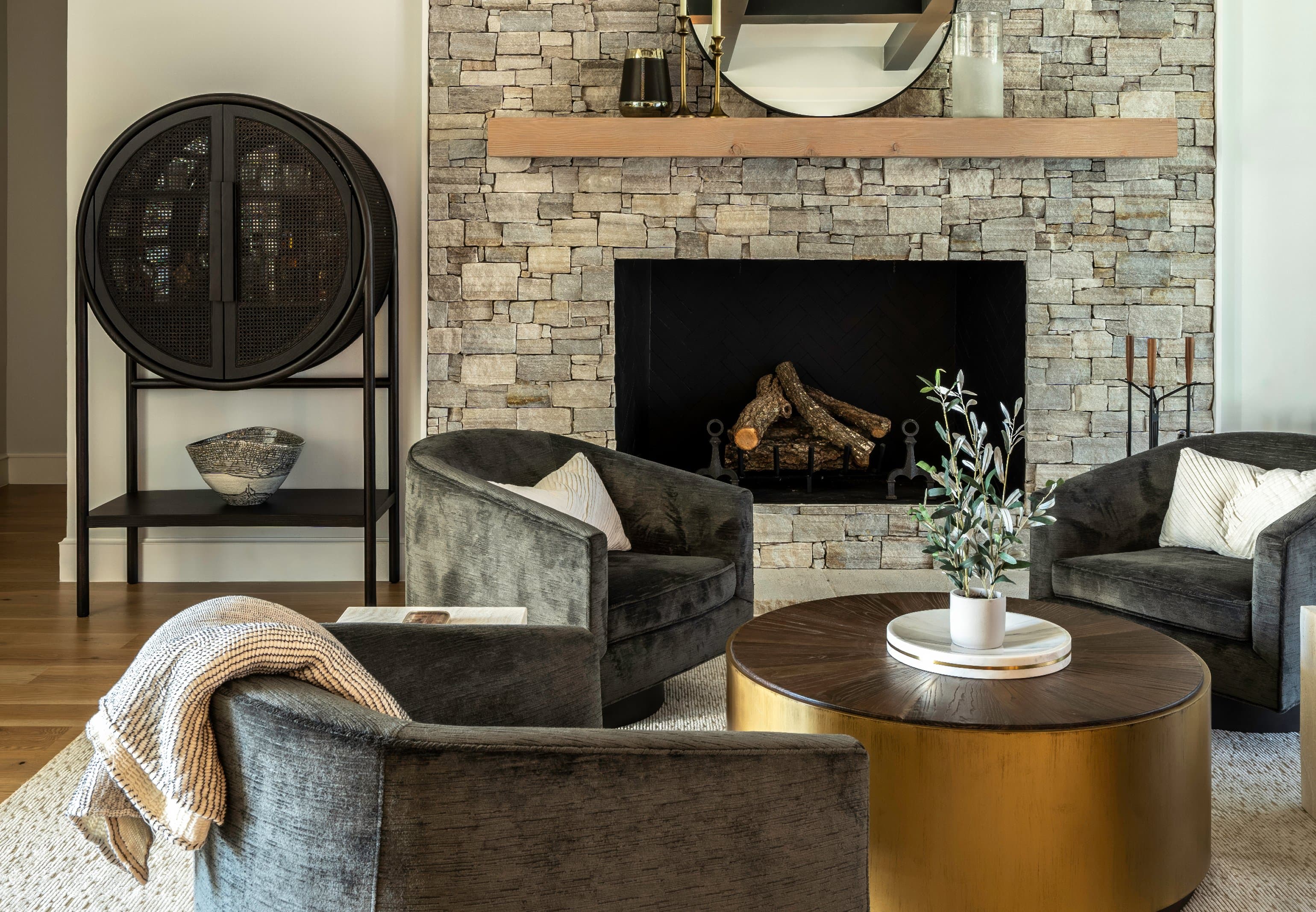 The Fairview sitting room: floor-to-ceiling stacked stone fireplace, charcoal velvet swivel chairs around a round brass coffee table, black bar cabinet on the left