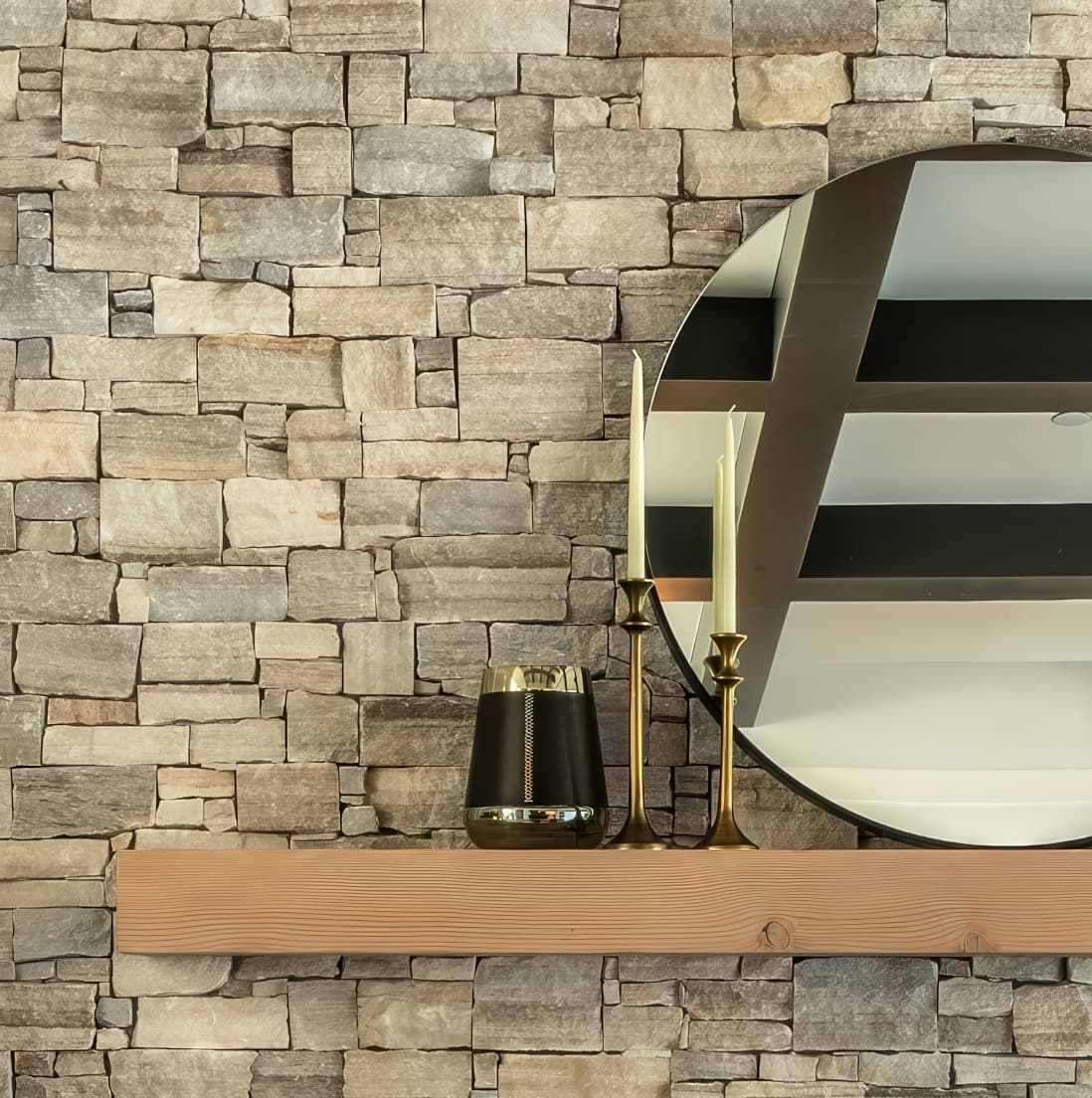 Detail of the stacked stone fireplace wall with wood beam mantel, round mirror, and brass candlesticks alongside a leather and brass tumbler