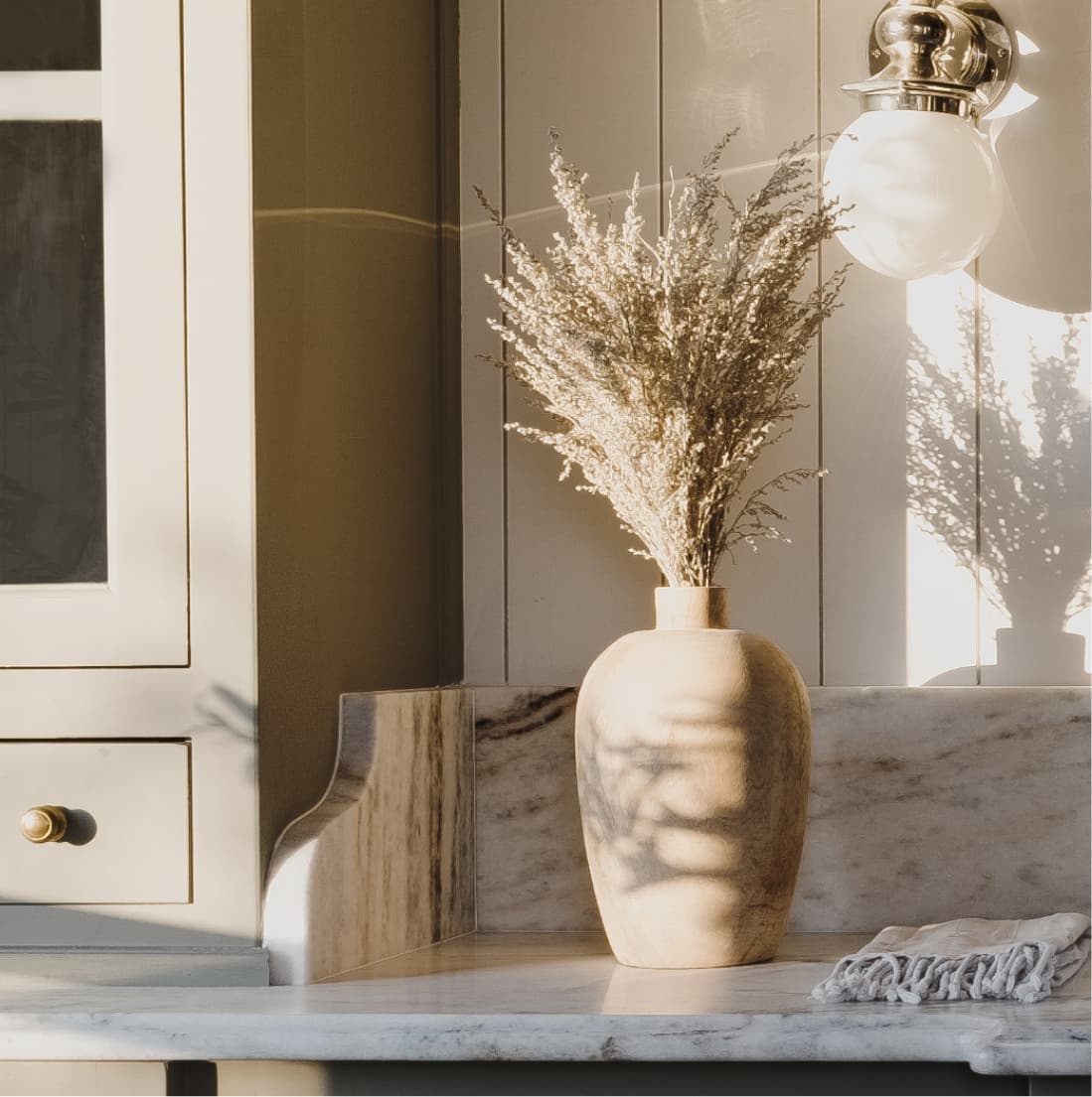 Detail, ceramic vase with dried botanicals on marble counter, globe sconce casting shadows