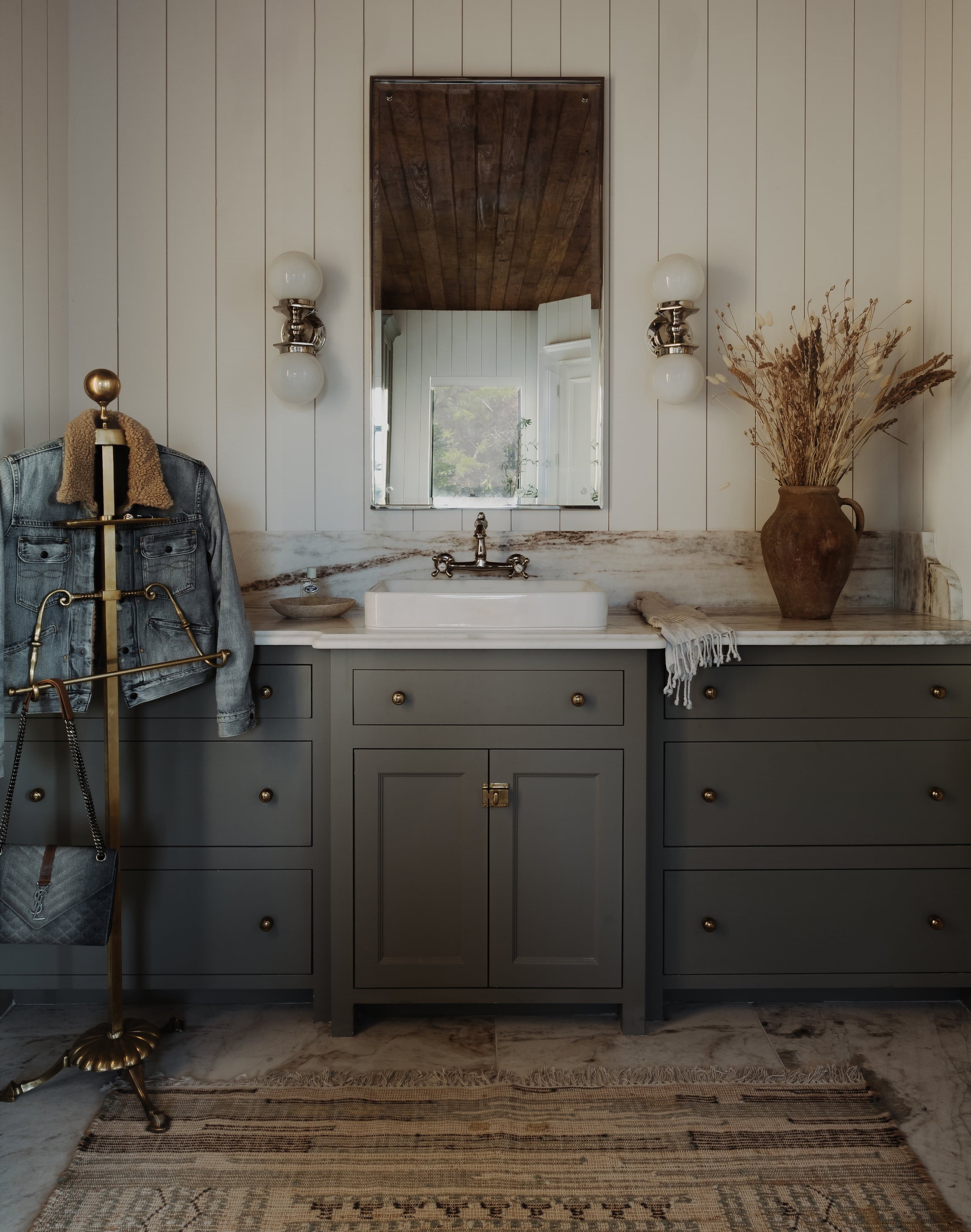 His vanity, brass valet stand with denim jacket, antique mirror, sage cabinetry, globe sconces