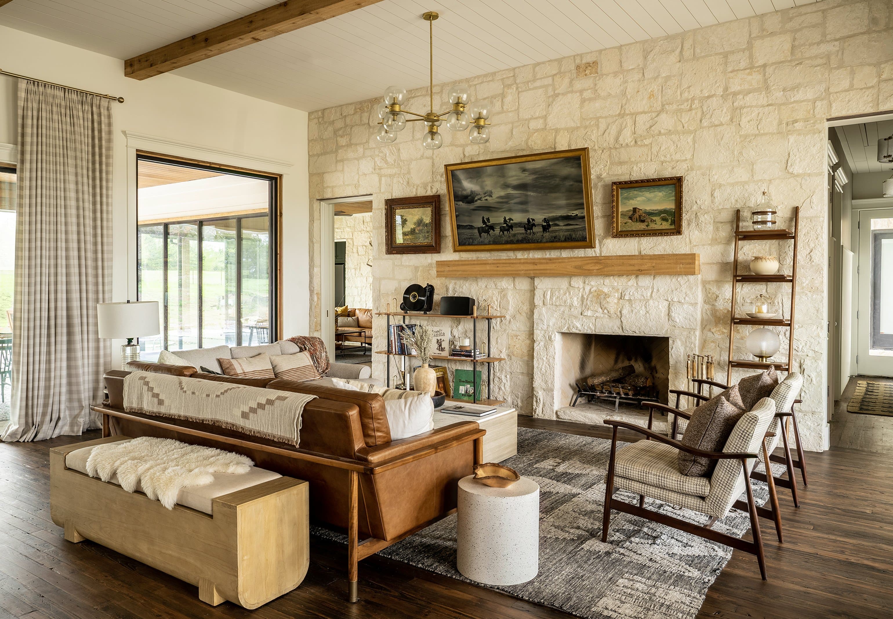 Hill Country Residence living room: floor-to-ceiling limestone fireplace wall, cognac leather sofa with Navajo-style throw, tweed armchairs, leather bench with sheepskin, brass pendant chandelier, reclaimed pine floors