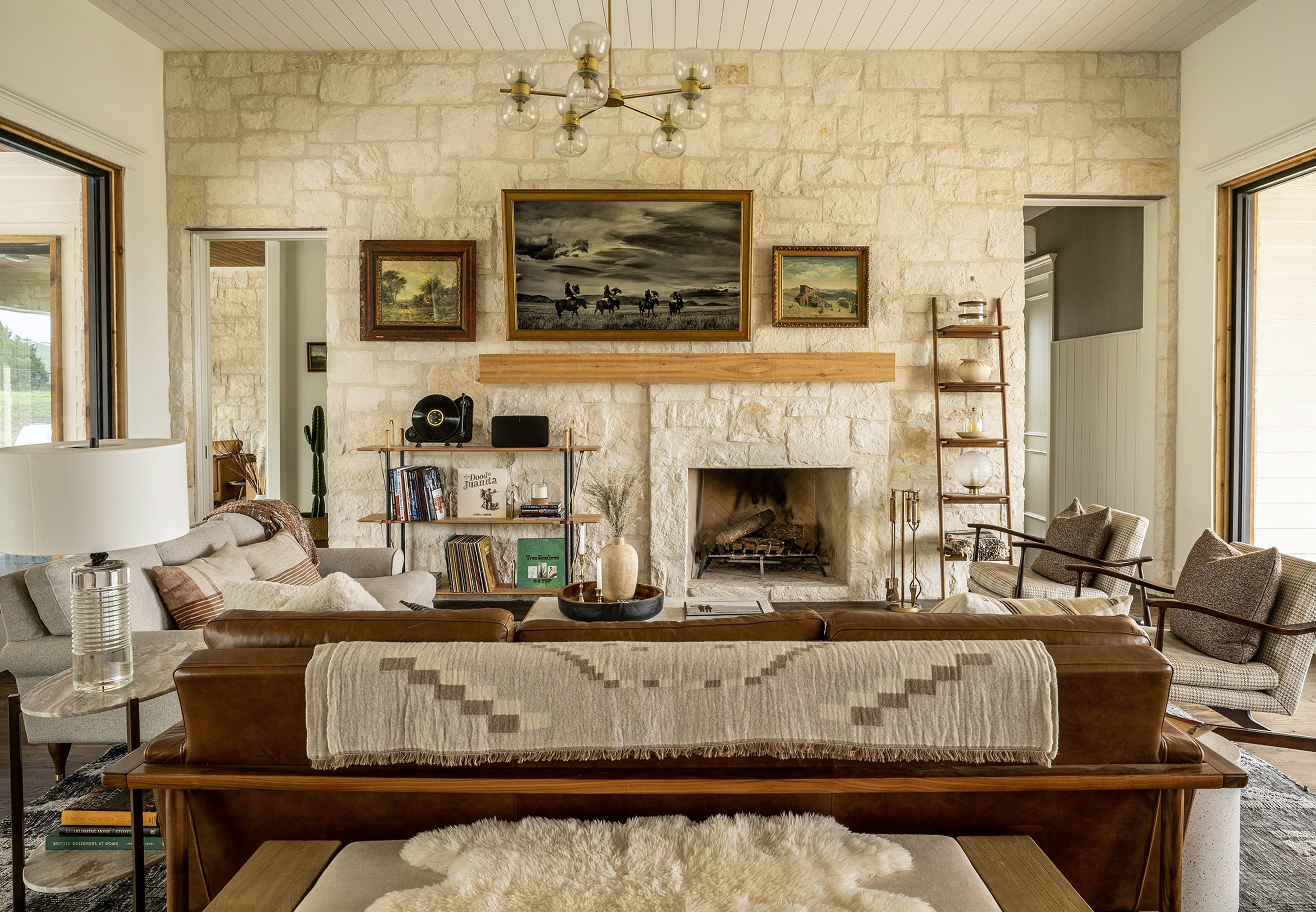 Symmetrical centered view of the limestone fireplace wall: Eisenhower painting flanked by landscape pieces, brass pendant chandelier overhead, cognac leather sofa with Navajo-style throw, every material in one frame
