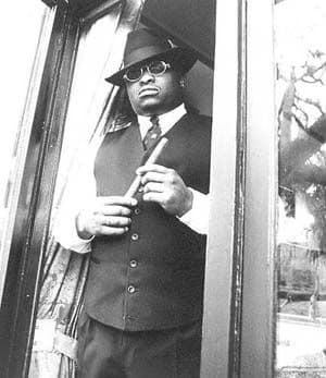 Black-and-white portrait, suited bluesman in fedora and round glasses with cigar in a doorway