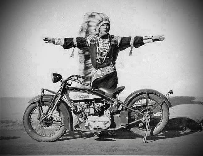 Black-and-white photograph, figure in feather headdress arms outstretched behind an Indian motorcycle
