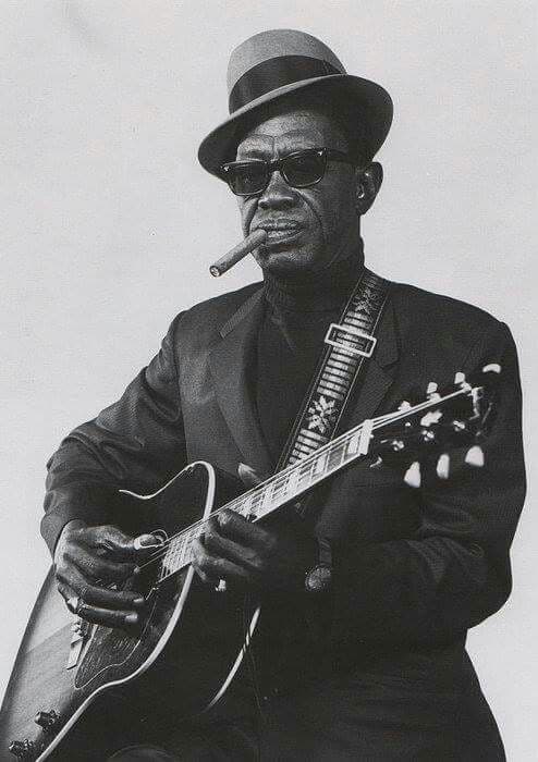 Black-and-white portrait of bluesman in fedora smoking a cigar with an archtop guitar
