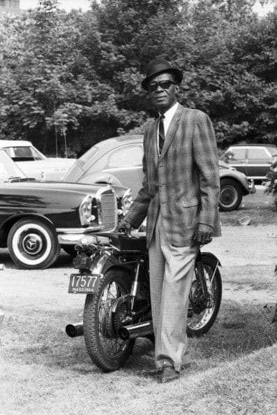 Black-and-white portrait, suited bluesman with a motorcycle and parked cars behind