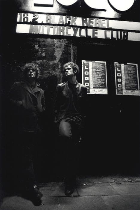 Black-and-white street shot of two musicians under a Black Rebel Motorcycle Club marquee