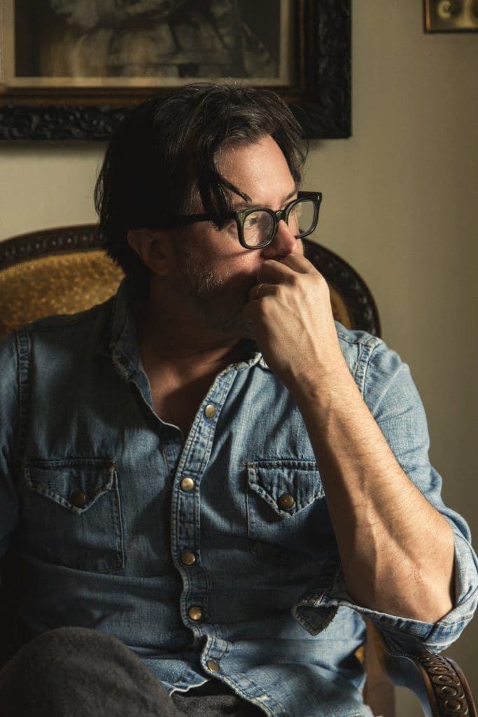 Bearded man in denim shirt and tortoiseshell glasses, seated in a carved armchair