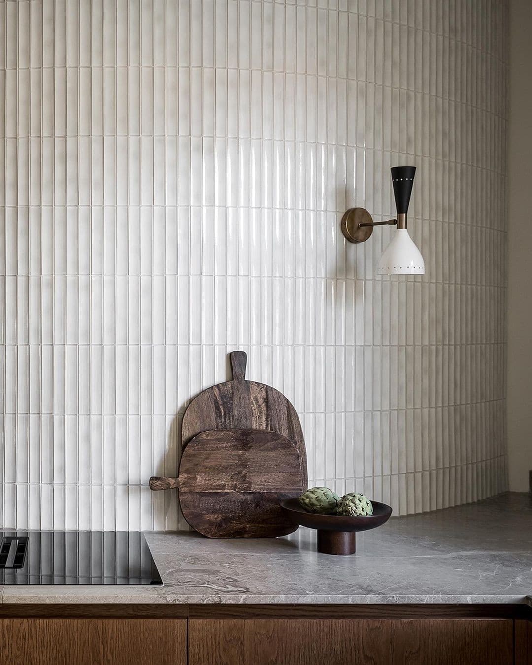 Modern kitchen with fluted white tile, stone counter, dark cutting boards, brass sconce