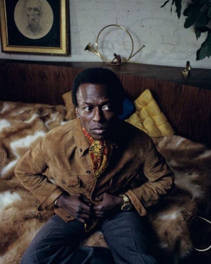 Miles Davis seated on fur bedspread in suede jacket and printed silk scarf
