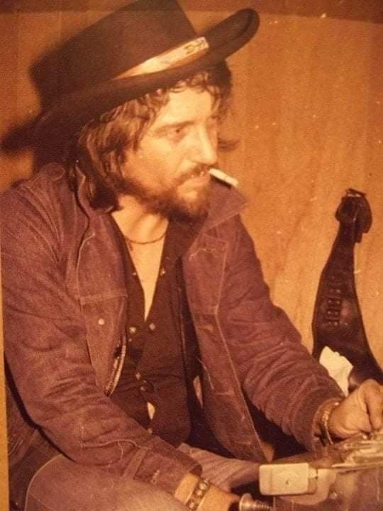 Sepia portrait of Waylon Jennings in cowboy hat smoking a cigarette beside a bottle