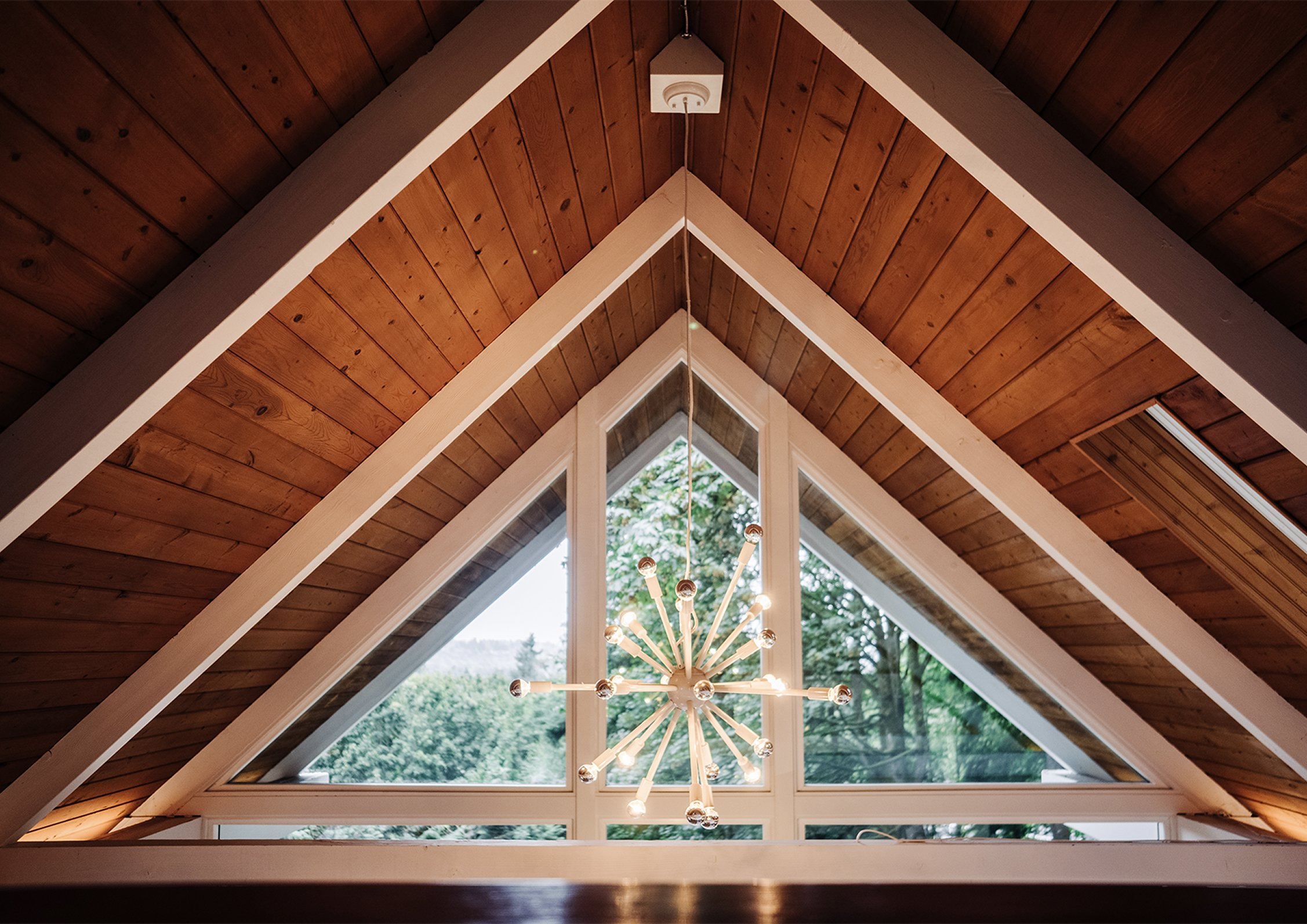 Looking up at the chalet's A-frame wood-plank ceiling with a sputnik chandelier suspended over a triangular window framing the tree canopy outside