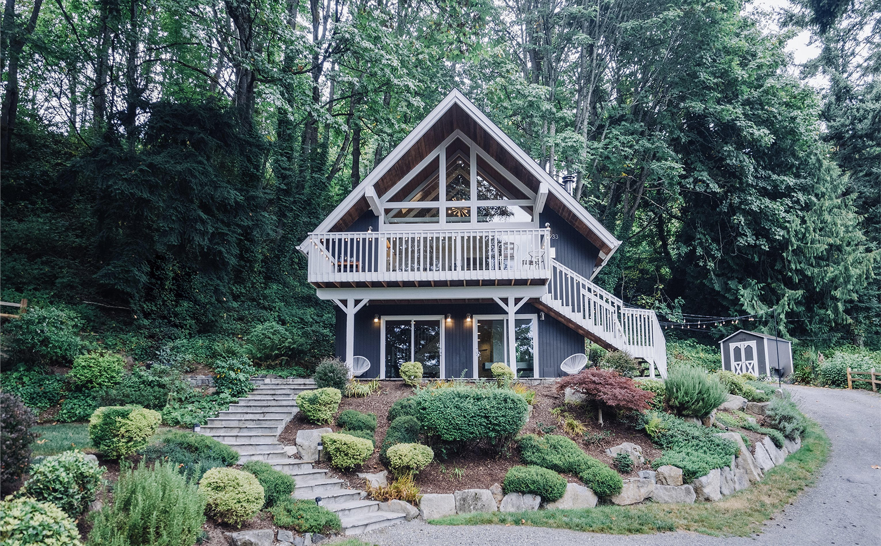 Front of the chalet repainted in warm gray with white railings, surrounded by Pacific Northwest evergreens and rocky landscape