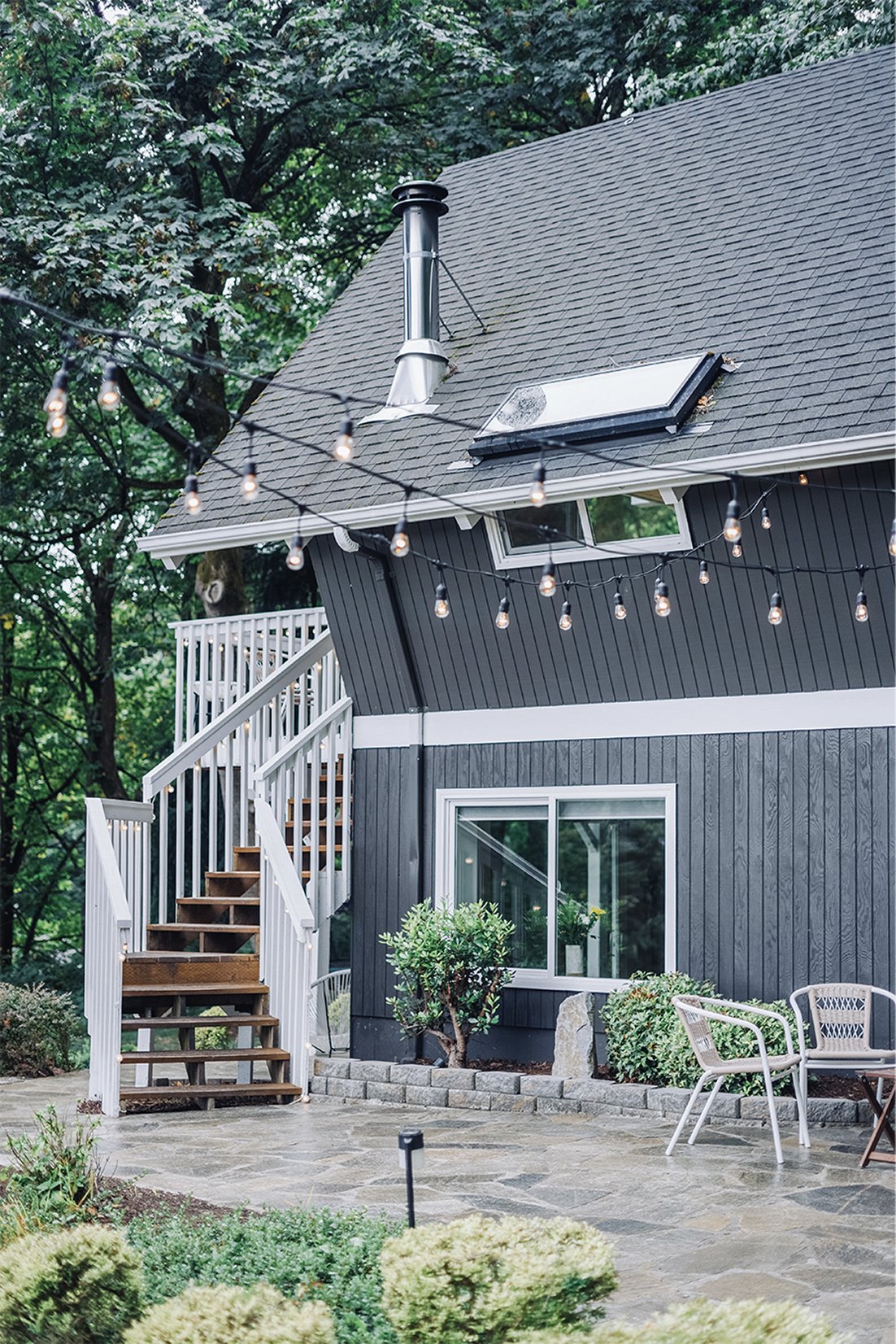 Side of the chalet at dusk with string lights strung over a stone patio, white-railed stairs, and warm gray siding