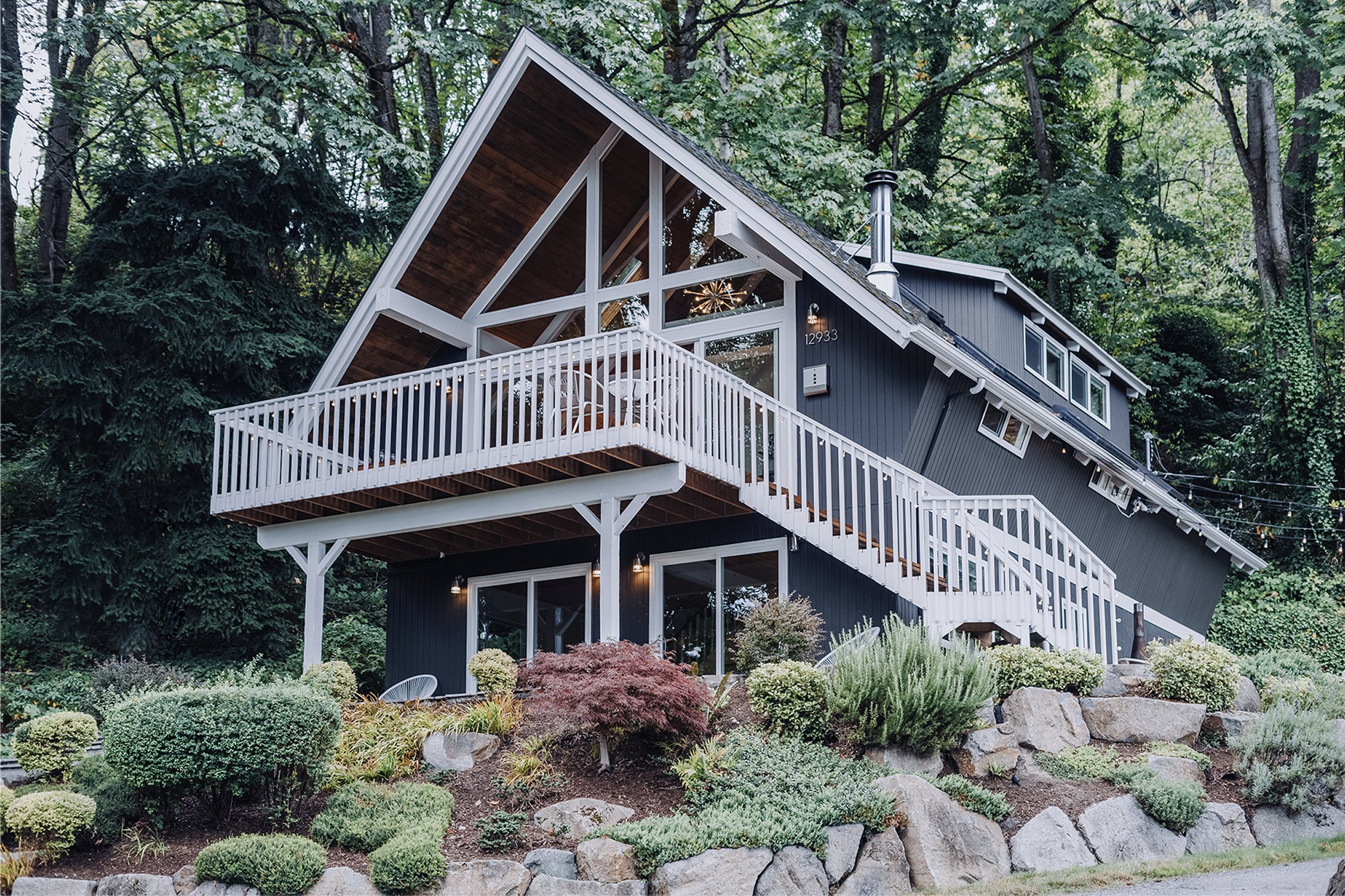 Three-quarter view of the chalet showing the deck, white-railed stairs, and the rocky landscape grade leading up to the front door