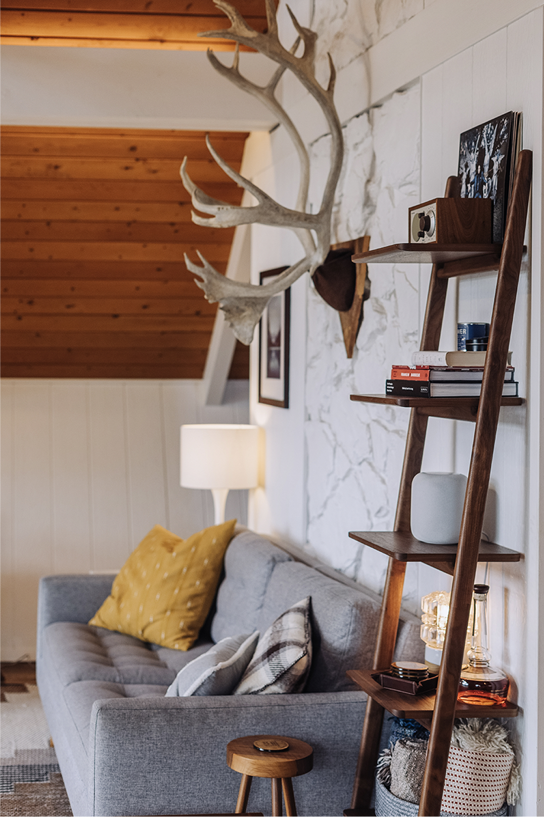 Detail of the living room with tufted gray sofa, walnut leaning ladder shelf, antler mount on a painted stone wall, and a yellow accent pillow