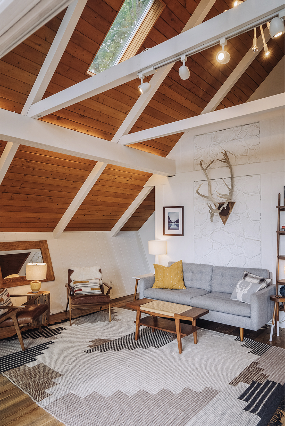 Wider chalet living room view with the A-frame ceiling, skylight, leather sling chair, walnut coffee table, and the painted stone wall in the background