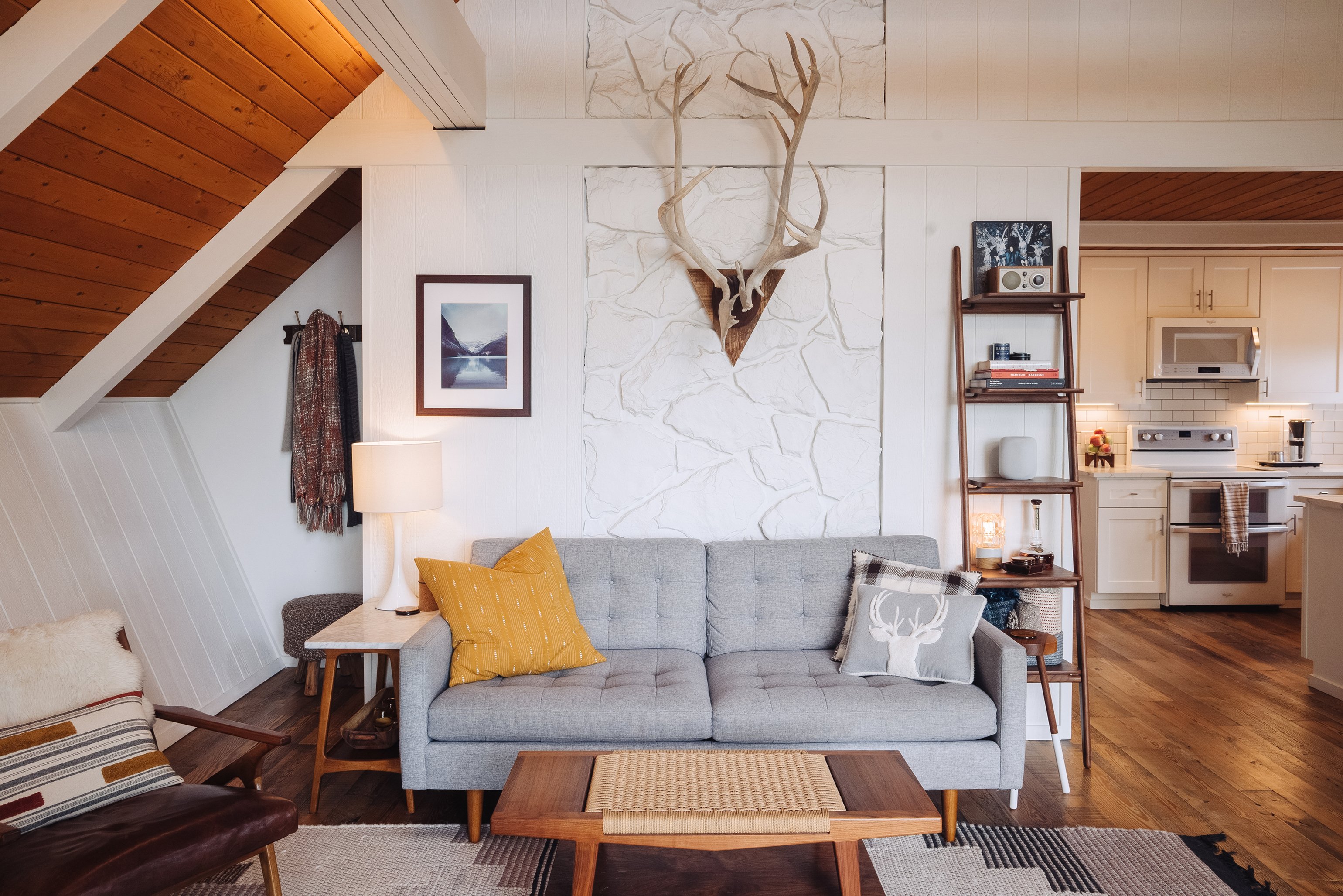 Chalet living area with tufted gray sofa, painted stone wall, antlers, walnut leaning ladder shelf, and the kitchen visible to the right
