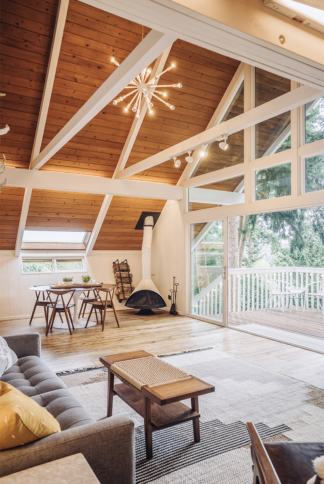 Wide view of the chalet living room with a walnut dining set, the Malm fireplace, sputnik chandelier overhead, and 16-foot glass doors opening to the deck