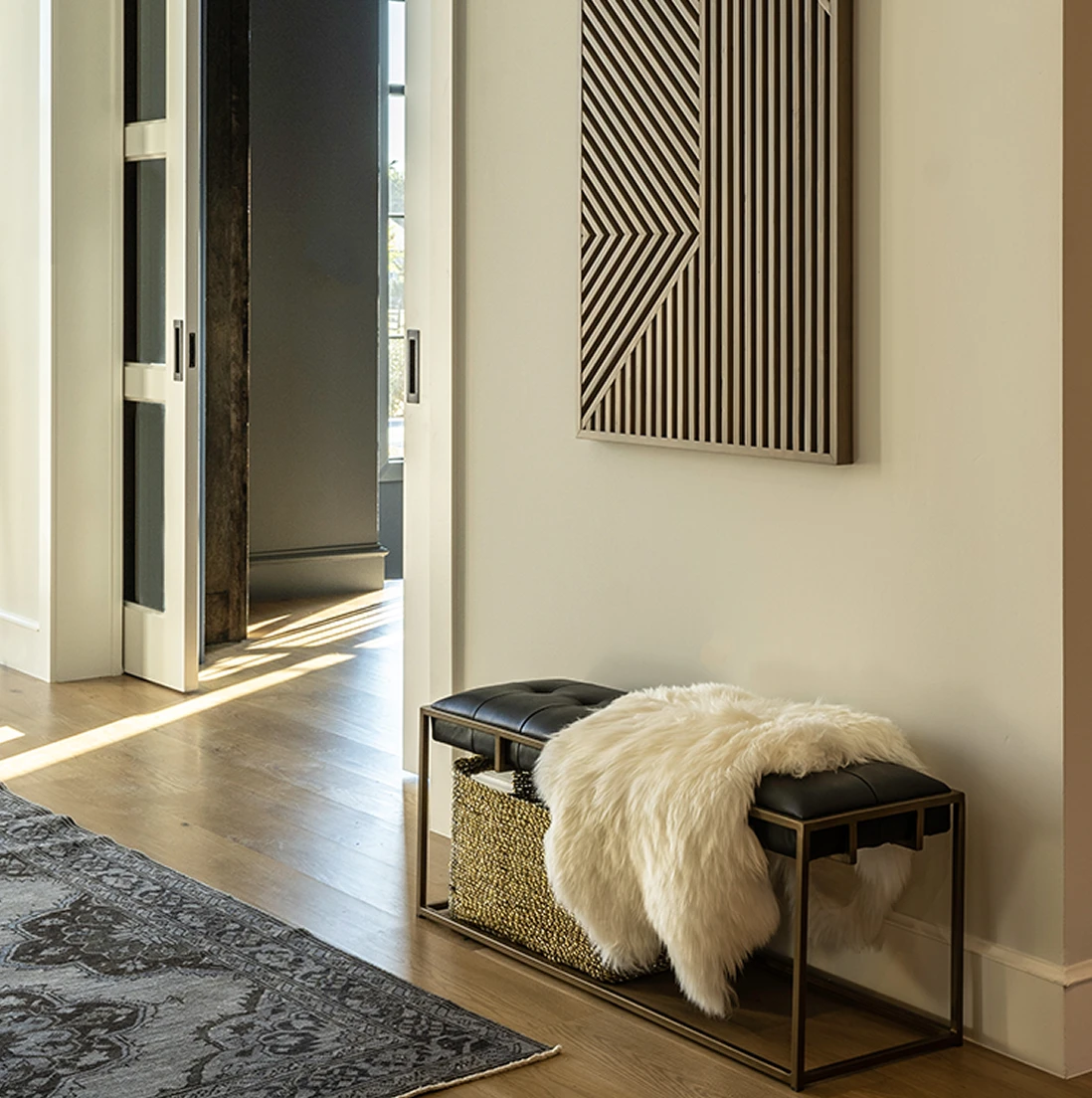 Tight detail of the black leather bench with a sheepskin throw, woven basket below, under the slatted wood geometric wall art