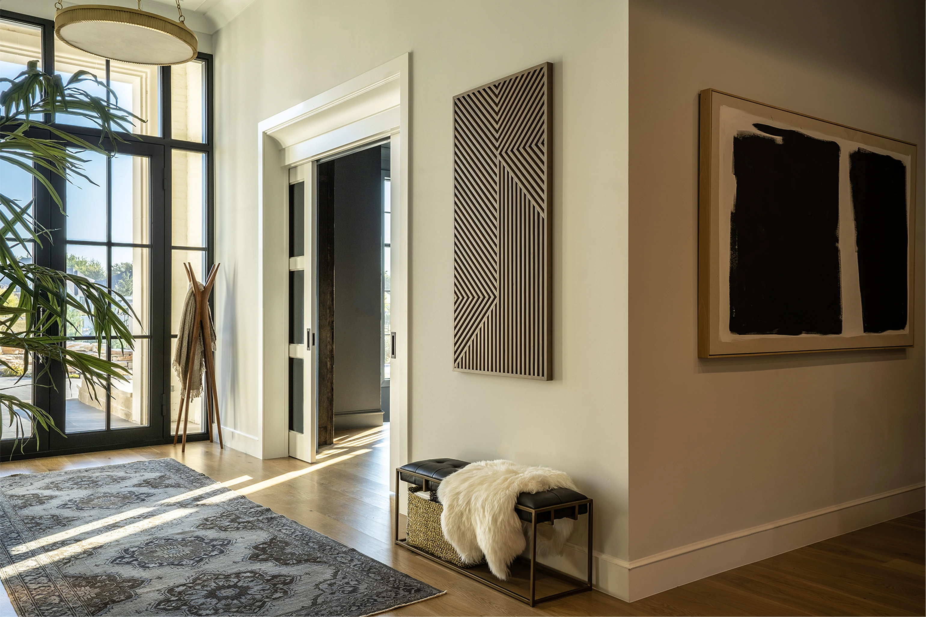 Side angle of the entry showing the full art wall: slatted wood geometric piece and dark abstract in a thick wood frame, leather bench beneath, coat tree and palm in the foreground