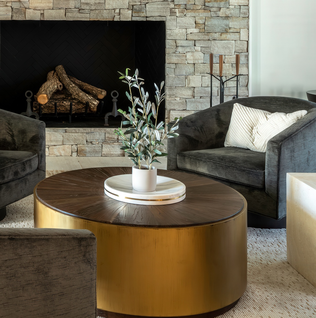 Tight detail of the round brass coffee table with wood top, olive plant in marble bowl, and the corner of a charcoal velvet swivel against the stone fireplace
