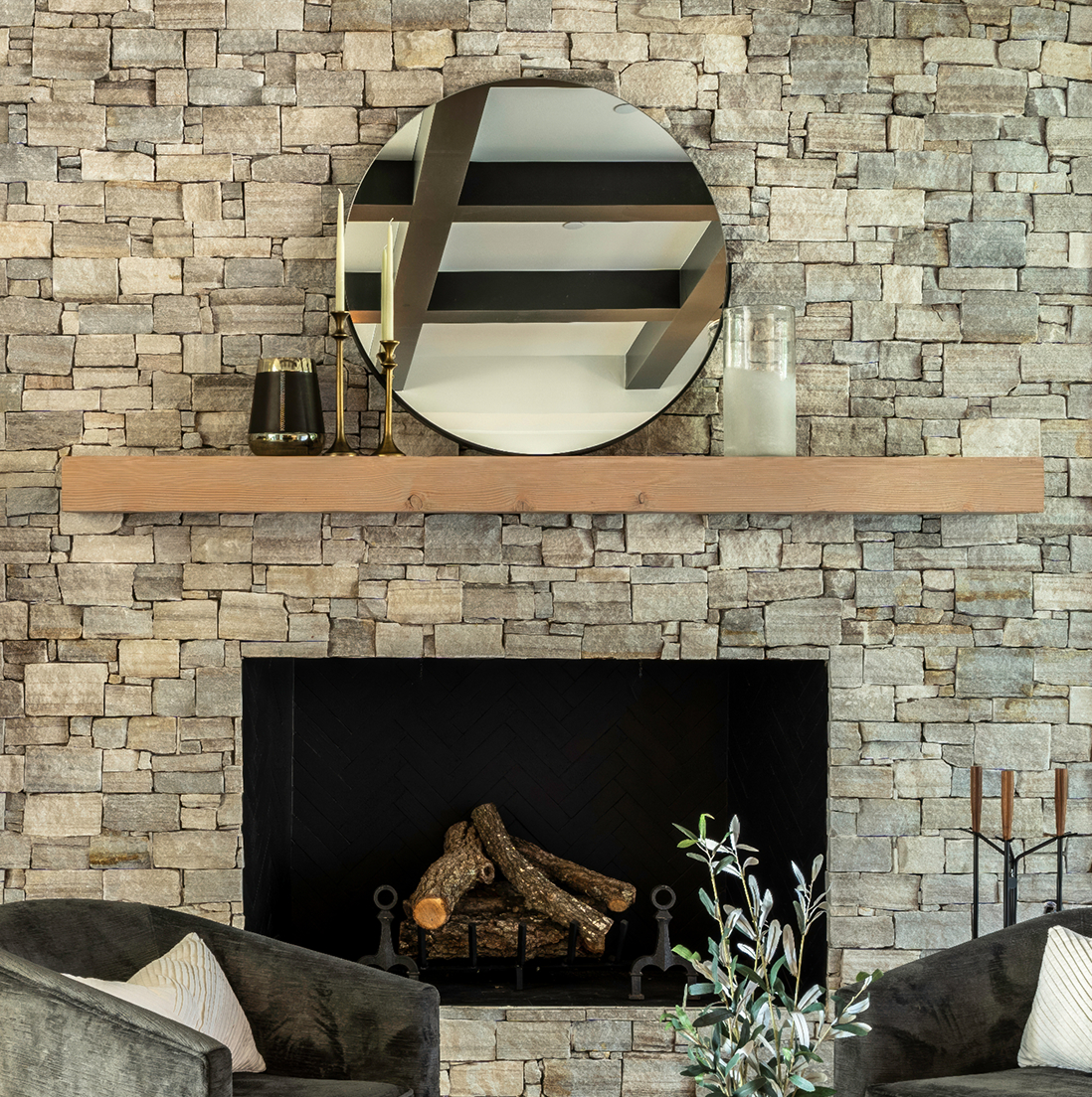 Centered shot of the wood mantel and round mirror against the stacked stone wall, with the fireplace and andirons below
