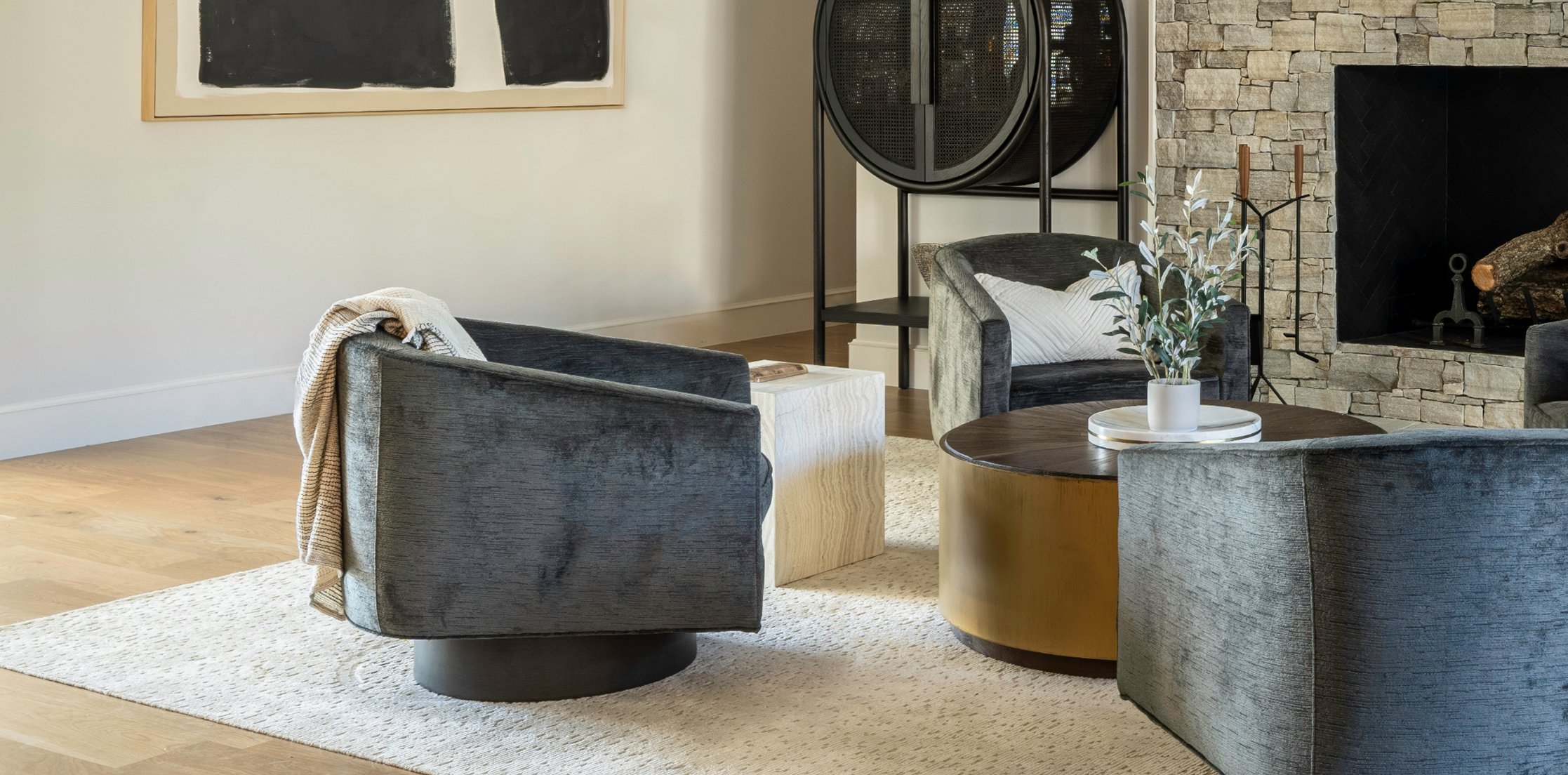 Rear view of the four charcoal velvet swivel chairs around the brass coffee table, throw blanket over one chair, large black abstract artwork on the wall behind, bar cabinet in view