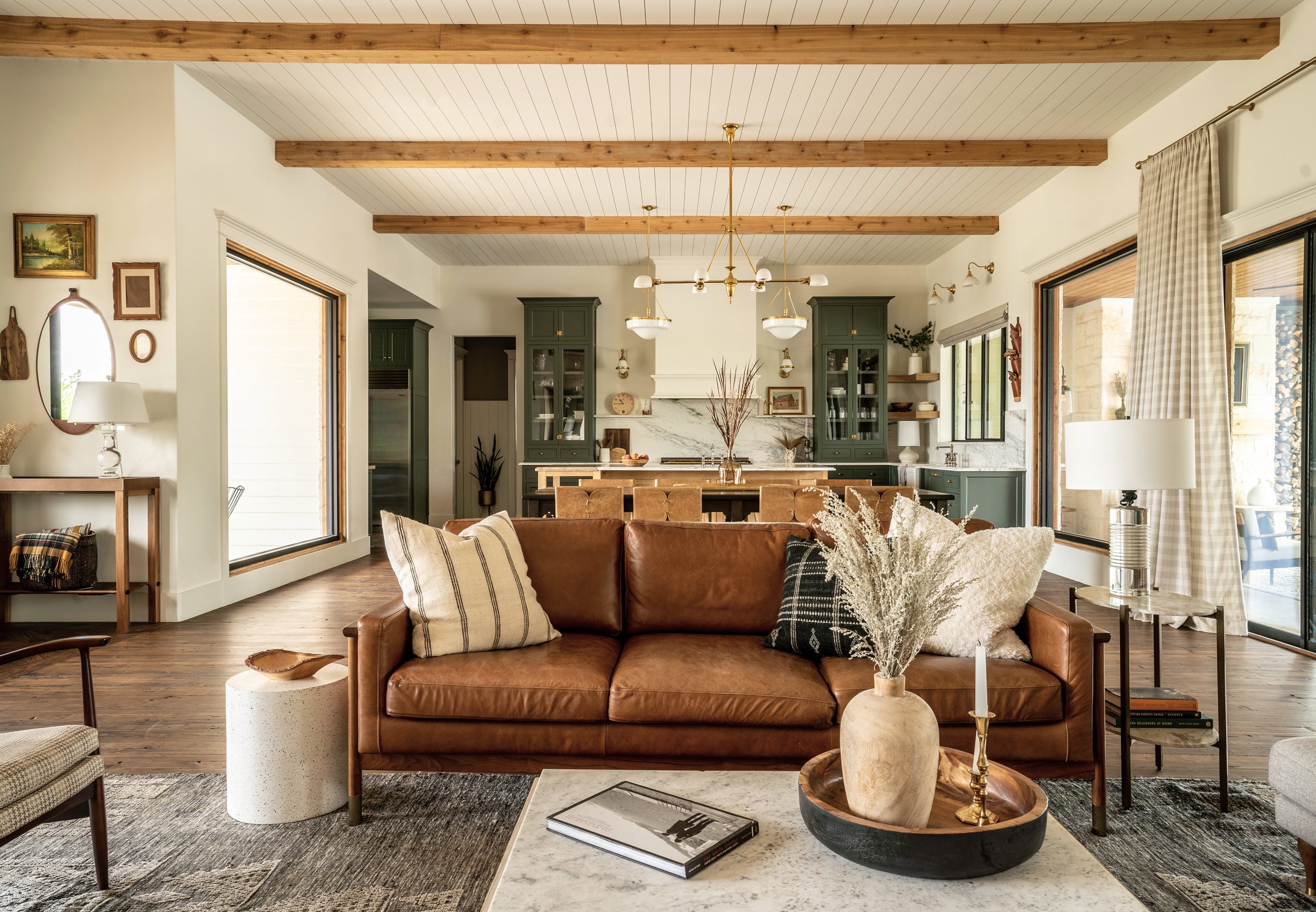 View from the living room into the open kitchen beyond: cognac leather sofa with pampas and textured pillows in the foreground, exposed wood beams overhead, green cabinetry and marble counters in the kitchen behind
