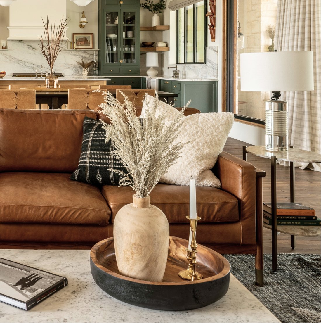 Tight detail of the cognac leather sofa with checkered pillow and pampas in a wood vase set in a black wood bowl on the coffee table, view through to the kitchen behind