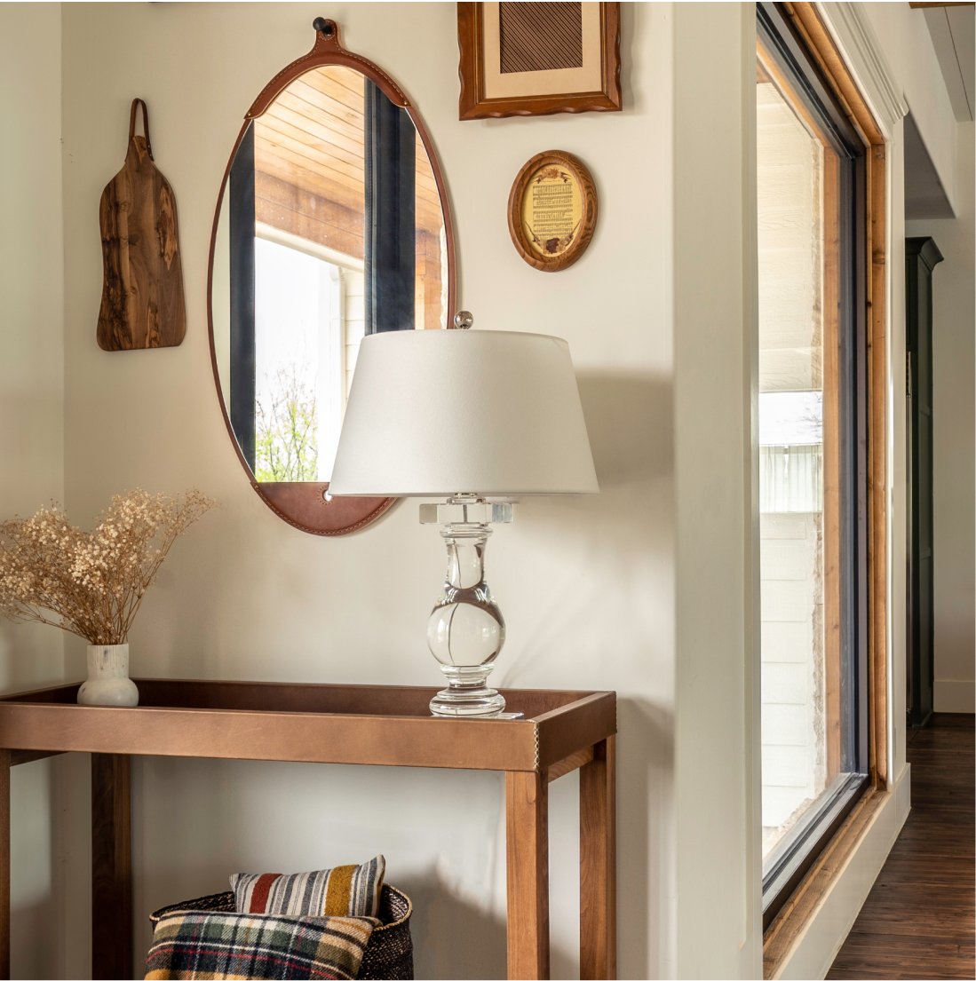 Entry vignette in an adjacent room: leather-strapped oval mirror, wood cutting board on the wall, small framed art, wood console table with crystal lamp, plaid throw and wicker basket beneath