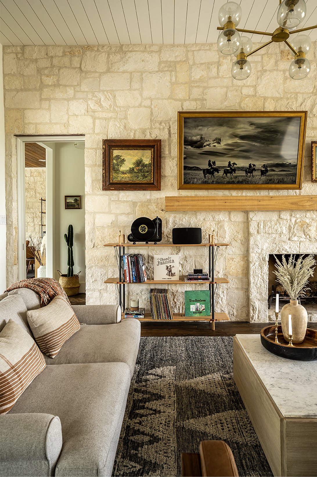 Vertical detail of the limestone wall, the Eisenhower painting and a smaller landscape framed in gilt, mid-century record-player shelf below, brass pendant chandelier overhead, edge of the gray sofa in the foreground