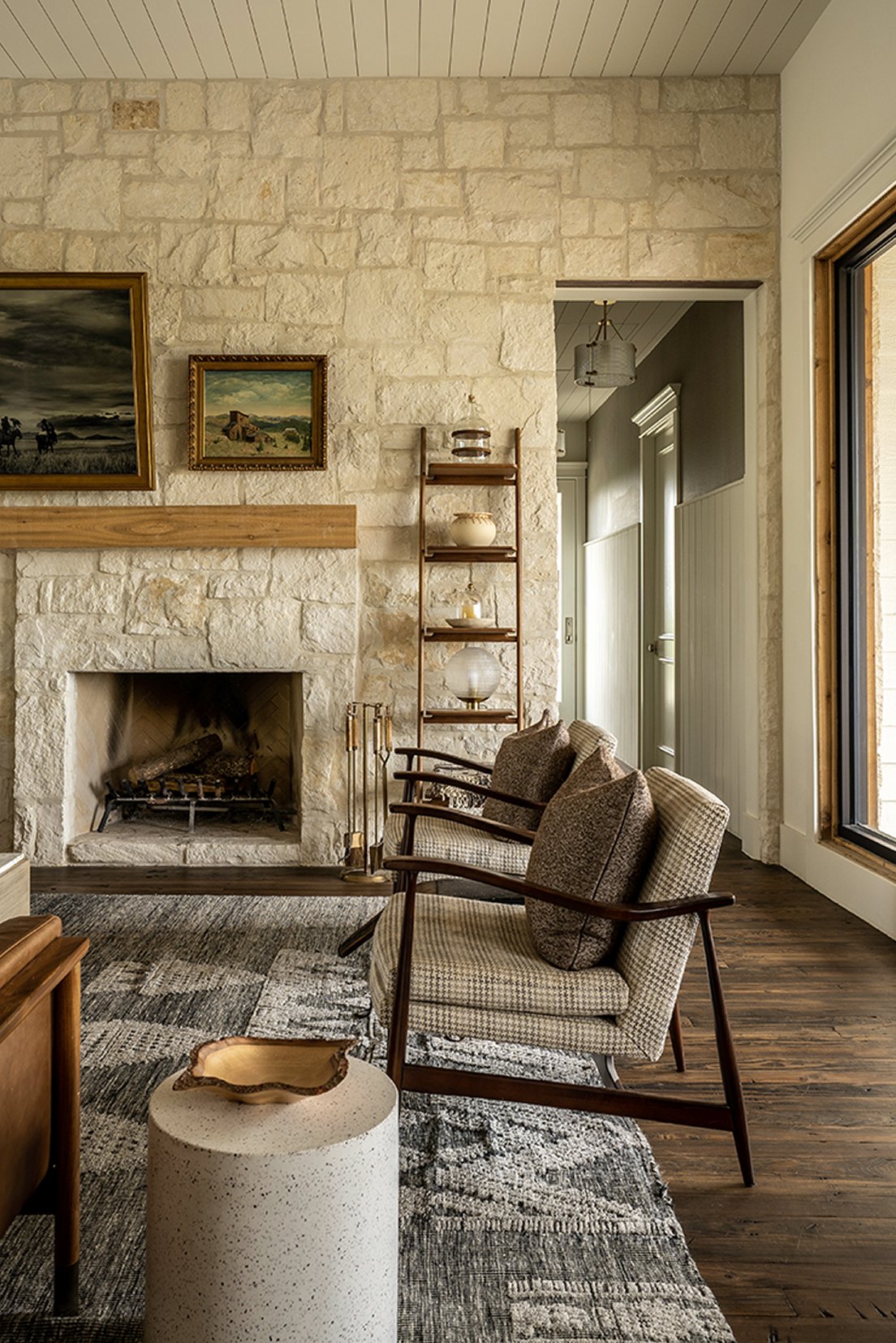 Vertical view of the limestone fireplace with the Eisenhower painting and a small landscape, tweed armchair beside it, ladder shelf with ceramic vessels, dark patterned vintage rug