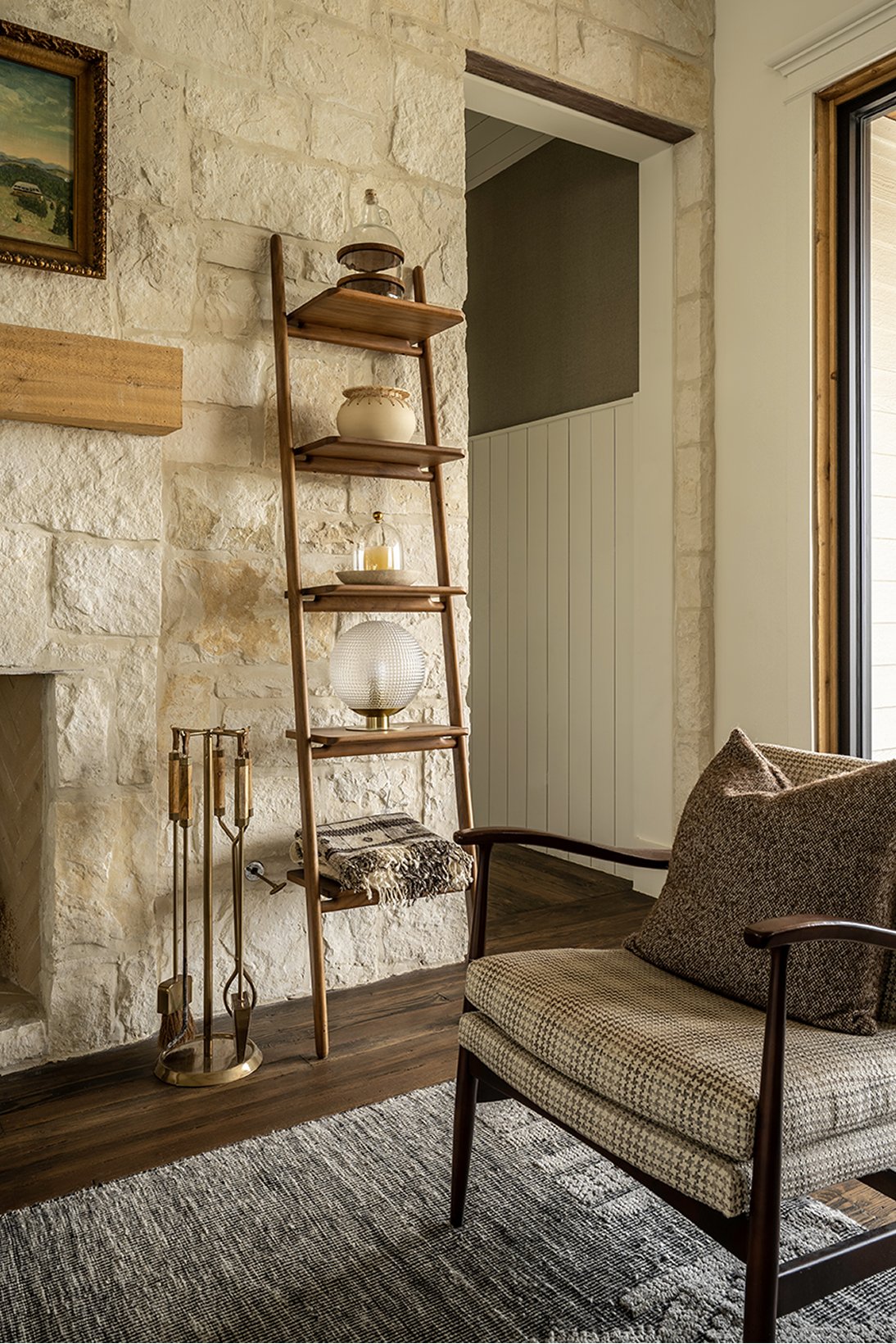Vertical detail of the limestone wall with the ladder shelf holding a vintage globe, brass fireplace tools below, and the corner of a tweed armchair