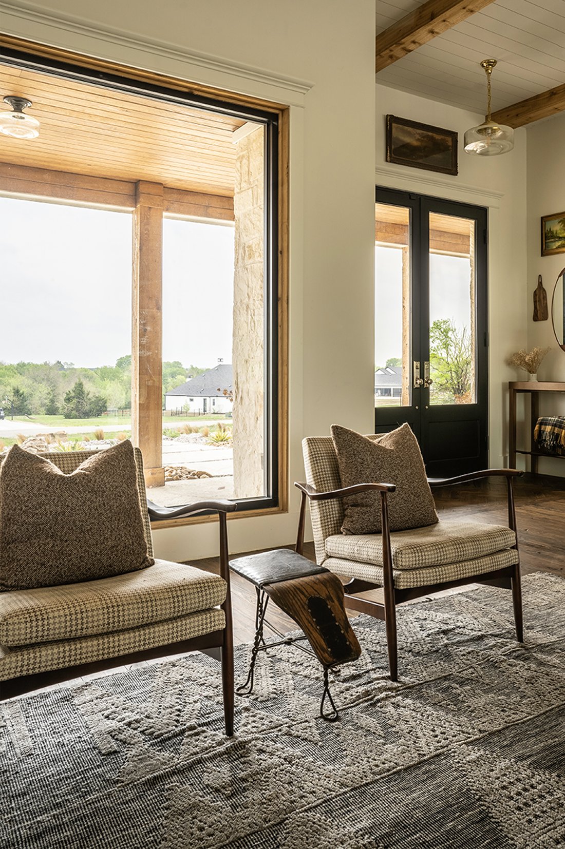 Two tweed armchairs facing each other in front of a tall window looking out to the Hill Country landscape, leather stool between them, vintage patterned rug underfoot