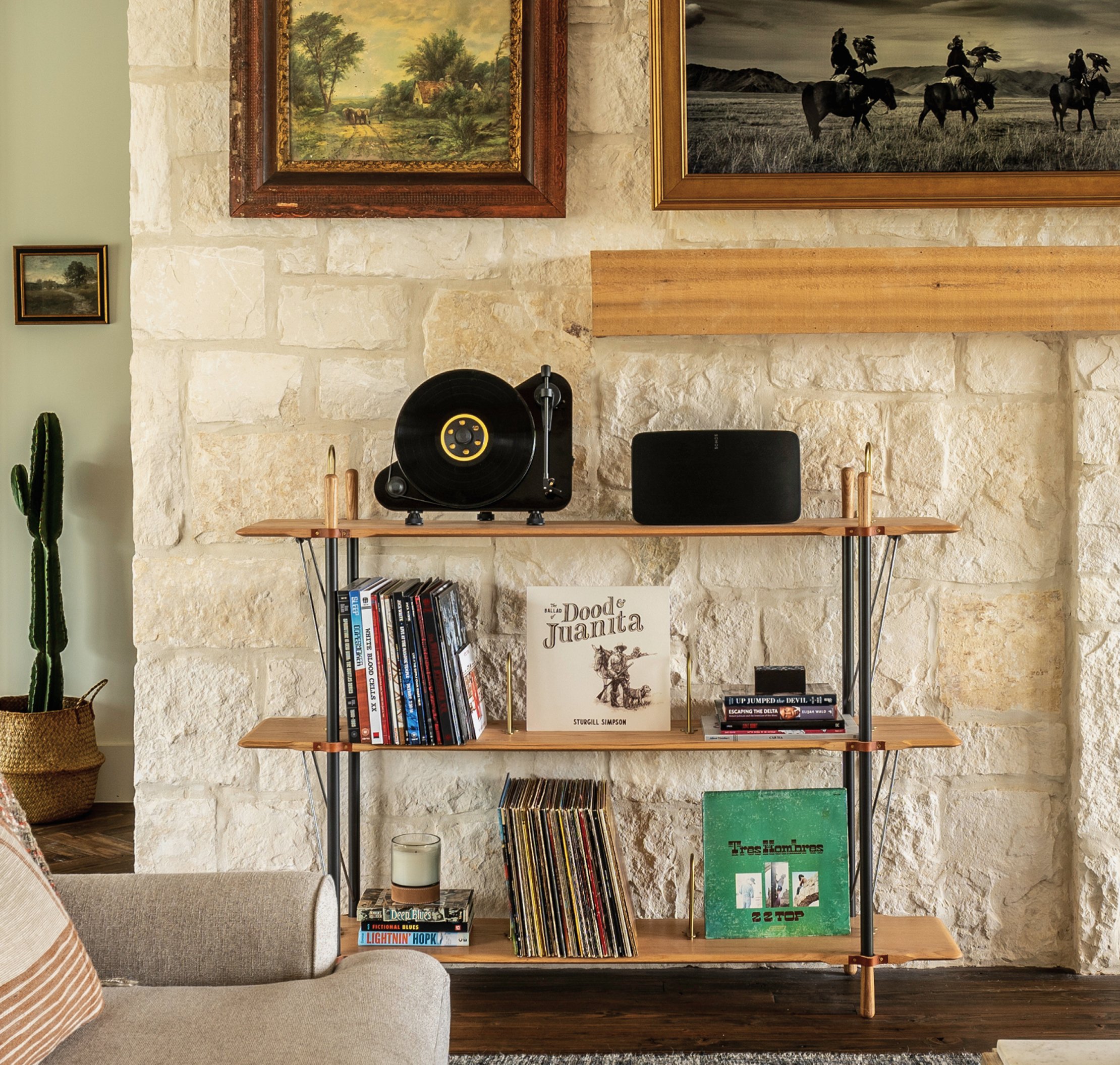 Tight detail of the mid-century record-player shelf against the limestone wall, vinyl records below including Sturgill Simpson and ZZ Top, cactus and small framed landscape painting to the side, edge of the Eisenhower painting visible above